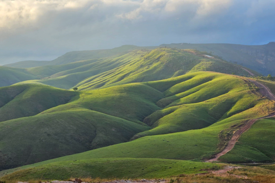 “Paisagem da Serra da Canastra com colinas verdes iluminadas pelo sol e estrada de terra sinuosa entre as montanhas.
