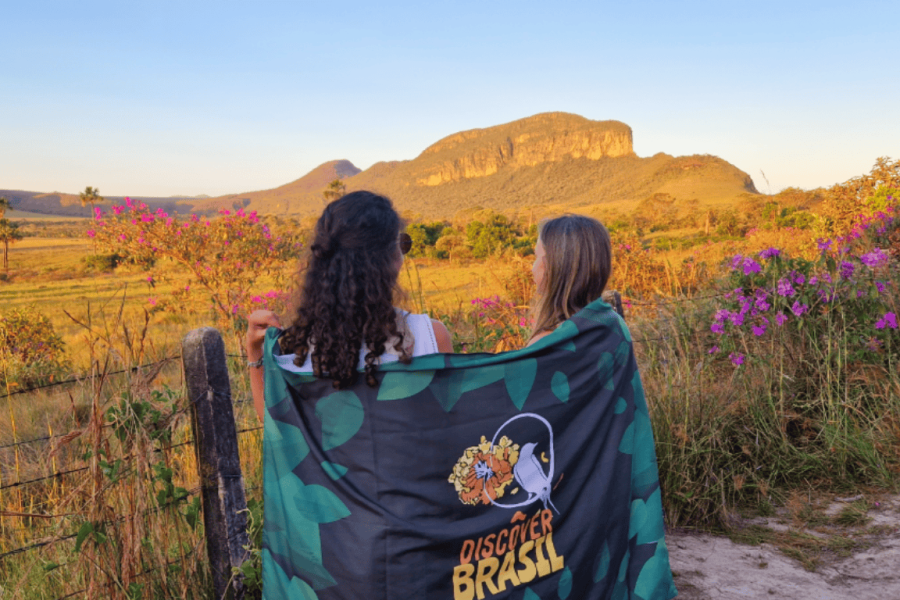 Two women admire the landscape of Jardim de Maytrea, in Chapada dos Veadeiros, surrounded by Cerrado wildflowers with the mountains in the background, during sunset.