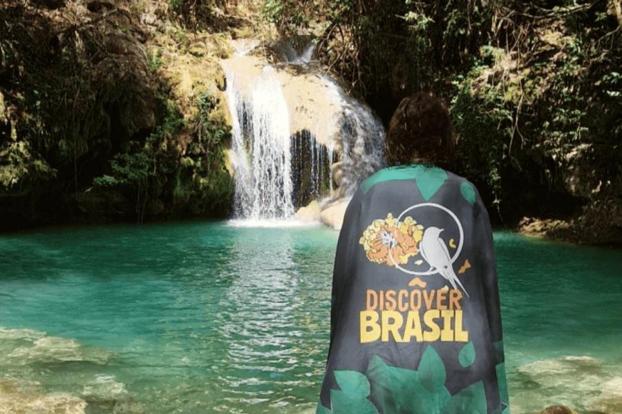 Person wearing a Discover Brasil cape admiring a turquoise waterfall in Mambaí, Goiás, surrounded by lush vegetation.
