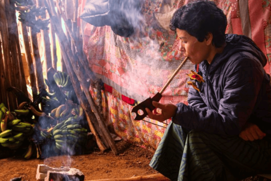 A Guarani Mbya Indigenous person during a ritual in the Tenondé Porã village, seated by the fire in a traditional setting, surrounded by cultural instruments and elements.