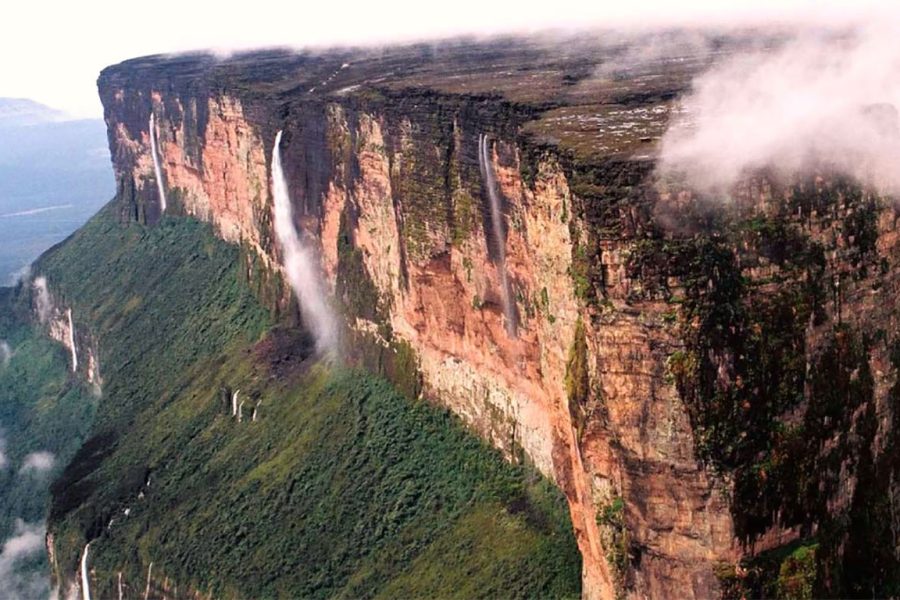 Aerial view of Mount Roraima with vertical cliffs, dense vegetation at the base, waterfalls cascading from the cliffs, and clouds surrounding the summit.