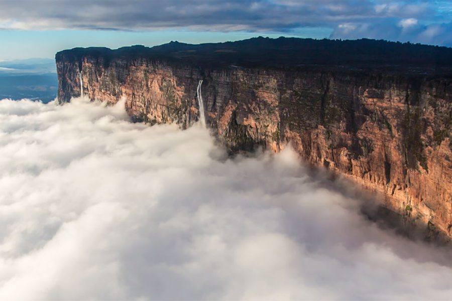 Aerial view of Mount Roraima surrounded by clouds, with a waterfall cascading down its rocky cliff on the border between Brazil, Venezuela, and Guyana.