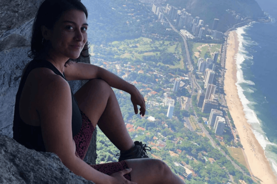 Woman sitting at the Garganta do Céu viewpoint, on Pedra da Gávea, with a panoramic view of Rio de Janeiro’s coastline and buildings in the background.