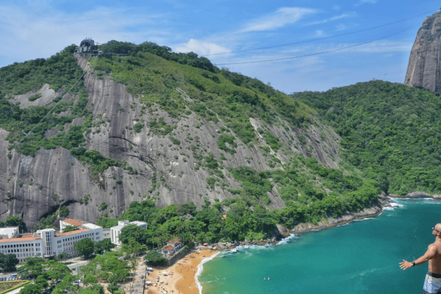 Panoramic view of Praia Vermelha in Rio de Janeiro, with hills covered in lush vegetation and Sugarloaf Mountain in the background, highlighting the natural beauty of the Babilônia Trail.