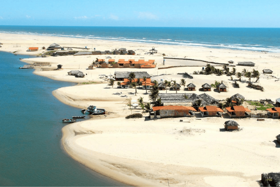 Aerial view of a coastal village in Lençóis Maranhenses, with simple houses among white sand dunes, a river meeting the sea, and the blue ocean in the background.